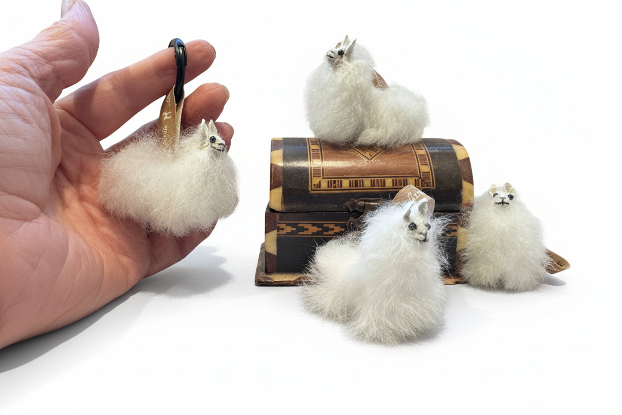 Four fluffy white llama or alpaca figurines with small faces, displayed on a dark wooden table with one held in a hand. One figurine is posed on top of an antique-style book.
