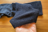 A close-up of a dark gray ribbed knit sock being held by a hand against a tan cardboard background.