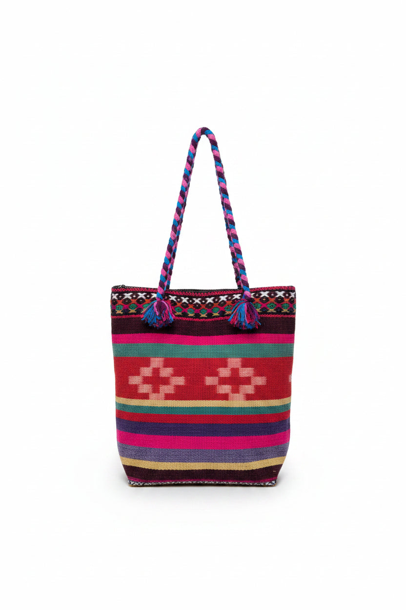 A colorful woven tote bag with braided handles, featuring a Southwestern geometric pattern in red, pink, purple, green, and beige, resting on a white shelf.
