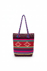 A colorful woven tote bag with braided handles, featuring a Southwestern geometric pattern in red, pink, purple, green, and beige, resting on a white shelf.