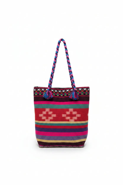 A colorful woven tote bag with braided handles, featuring a Southwestern geometric pattern in red, pink, purple, green, and beige, resting on a white shelf.