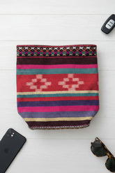 A colorful woven tote bag with braided pink and blue handles, resting on a light wood table next to a notebook and glasses.