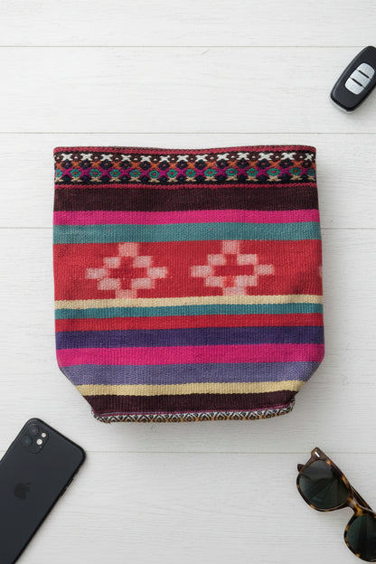 A colorful woven tote bag with braided pink and blue handles, resting on a light wood table next to a notebook and glasses.
