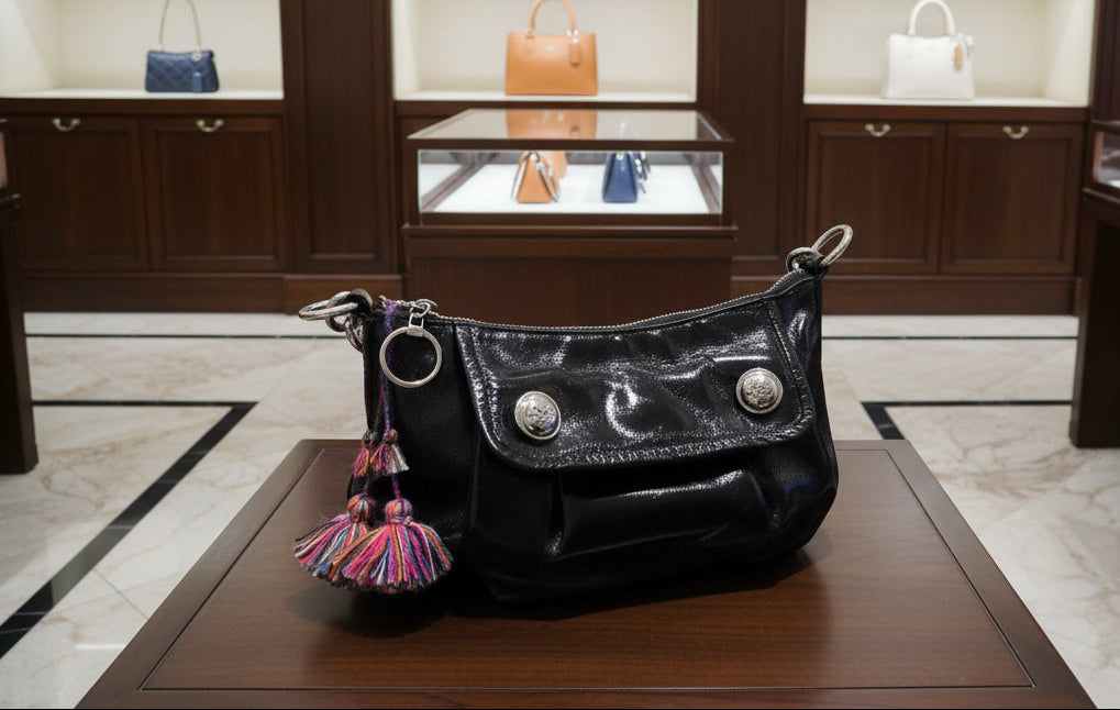 A black patent leather shoulder bag with silver-tone hardware, featuring a flap closure with decorative silver studs and colorful fringe tassels hanging from the side rings, shown resting on a wooden table.