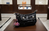 A black patent leather shoulder bag with silver-tone hardware, featuring a flap closure with decorative silver studs and colorful fringe tassels hanging from the side rings, shown resting on a wooden table.