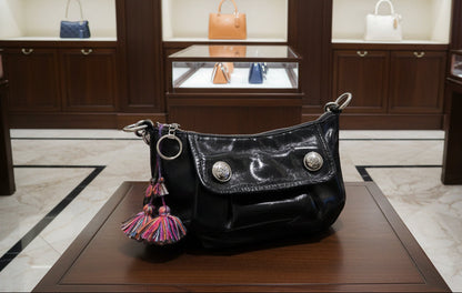 A black patent leather shoulder bag with silver-tone hardware, featuring a flap closure with decorative silver studs and colorful fringe tassels hanging from the side rings, shown resting on a wooden table.