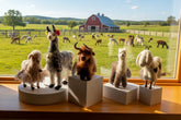 A set of five handcrafted felt animal figurines displayed on white pedestals against a white studio background. The collection includes a fluffy beige sheep, a gray and white llama with a red flower, a brown highland cow, a white and brown alpaca, and a white goat with black spots.