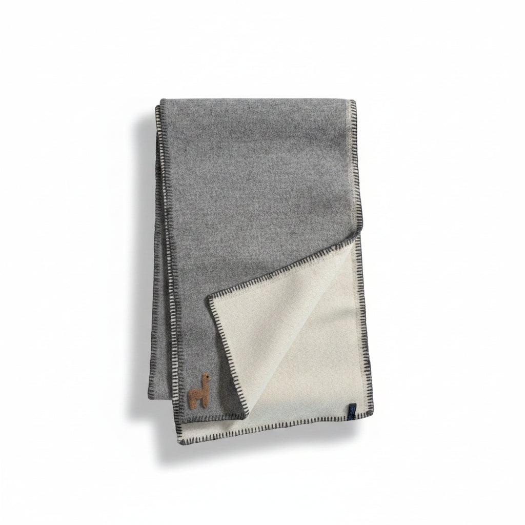 A folded gray wool blanket with a cream-colored reverse side and a small embroidered camel logo, displayed on a wooden floor.
