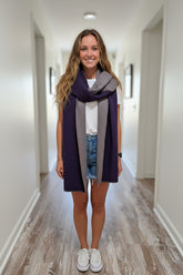 A smiling woman stands indoors wearing a folded dark purple and heather gray knit scarf or wrap around her shoulders, paired with a white t-shirt, denim shorts, and white sneakers.