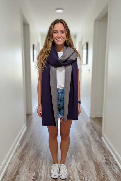 A smiling woman stands indoors wearing a folded dark purple and heather gray knit scarf or wrap around her shoulders, paired with a white t-shirt, denim shorts, and white sneakers.