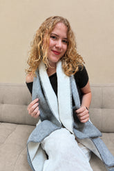 A woman with curly blonde hair sits on a sofa wearing a gray wrap-style scarf. She is holding the edges of the scarf to show both sides.