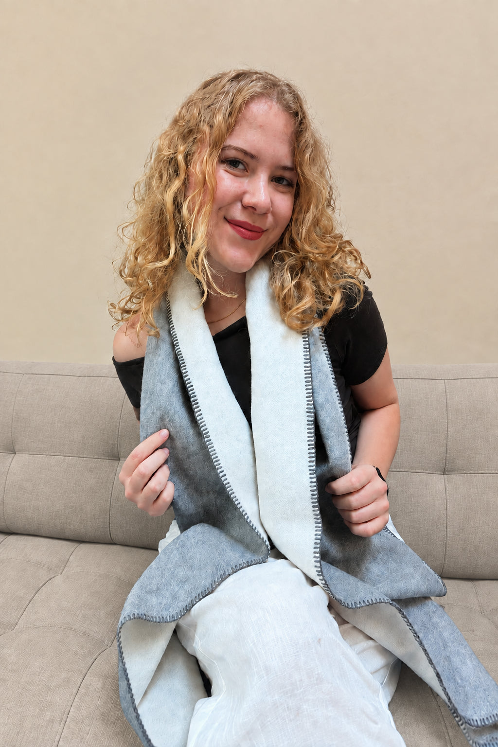 A woman with curly blonde hair sits on a sofa wearing a gray wrap-style scarf. She is holding the edges of the scarf to show both sides.