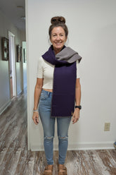 A smiling woman stands indoors wearing a folded dark purple and heather gray knit scarf or wrap around her shoulders, paired with a white t-shirt, denim shorts, and white sneakers.
