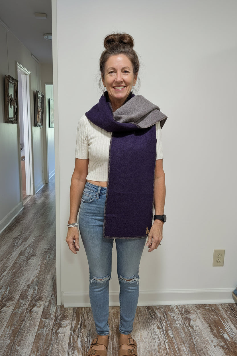 A smiling woman stands indoors wearing a folded dark purple and heather gray knit scarf or wrap around her shoulders, paired with a white t-shirt, denim shorts, and white sneakers.