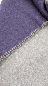 A close-up of a soft-woven reversible scarf, showing two folded sections: one in muted purple with a tan zigzag stitched edge, and one in light beige. The fabric texture is clearly visible.