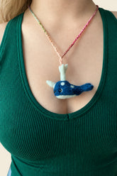 A close-up of a person wearing a handmade felt whale pendant necklace on a pink chain, paired with a green ribbed tank top.