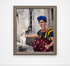 A framed portrait painting of Frida Kahlo with a white alpaca, displayed on a wooden floor against a white wall. The painting features Frida in her signature blue flower hair accessory, yellow and blue dress, and red skirt, reaching toward the white alpaca.