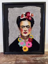 A framed portrait artwork of Frida Kahlo, featuring textured wool or felt-like fibers, a black frame, and a black-and-white background, displayed on a wooden surface against a white wall.