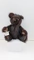 A dark brown shaggy teddy bear sitting on a white display block, with a small paper tag attached to its front paw.