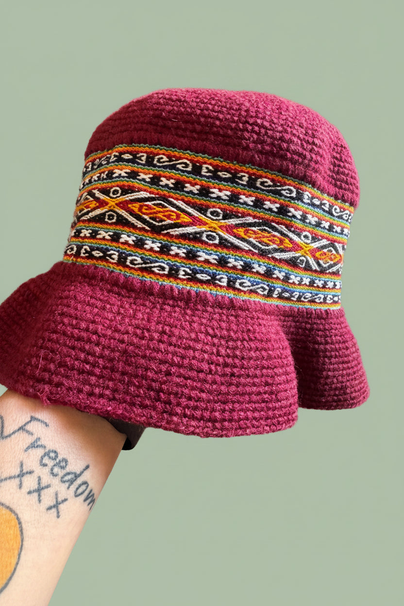A close-up of a hand-crocheted bucket hat in deep magenta, featuring a wide decorative band with geometric patterns in orange, yellow, green, and white. The hat is shown resting on a striped cushion with a stone tile floor in the background.