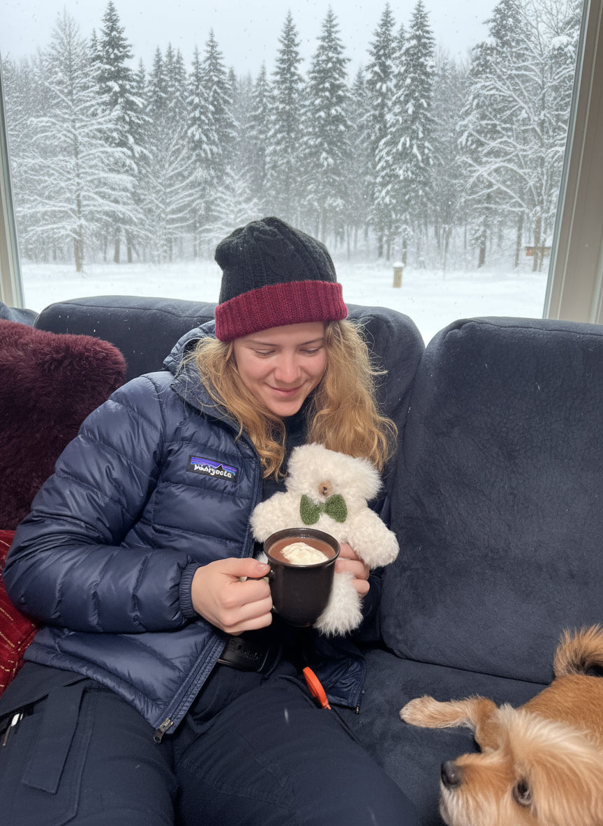 A person sitting on a couch with an Irish reversible beanie and dog and a teddy bear, holding a mug in a snowy outdoor setting.