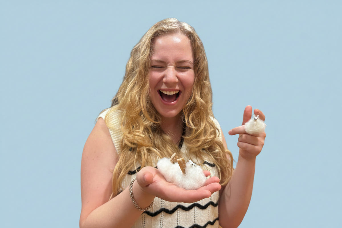 A smiling woman with long blonde hair holds a small white fluffy bird in her hands inside a home, with white cabinets and a living room visible in the background.