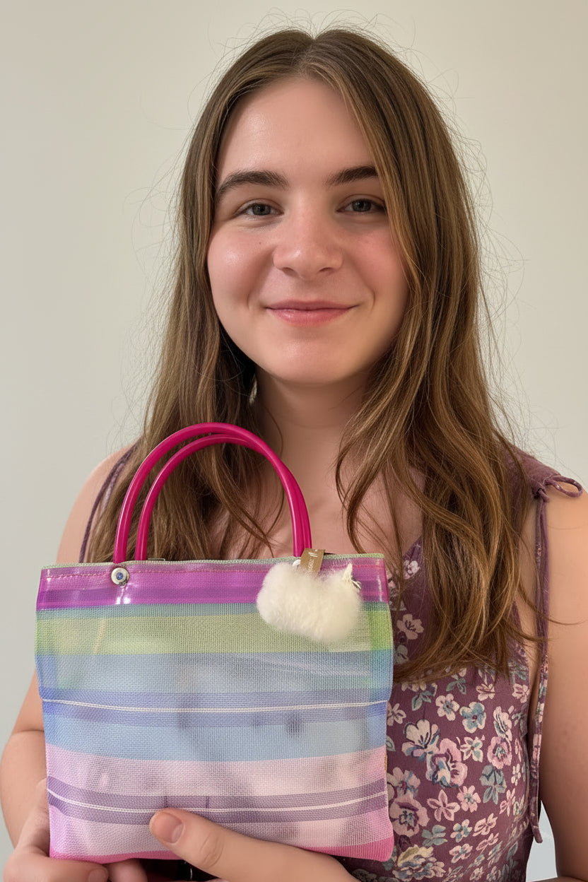 A young woman holding a translucent striped tote bag with pink handles and a white pom-pom charm, wearing a floral top against a plain light background.
