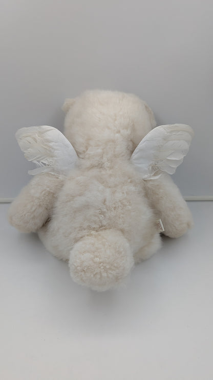 A fluffy cream-colored teddy bear plush toy with white angel wings, photographed from behind against a plain light gray background.