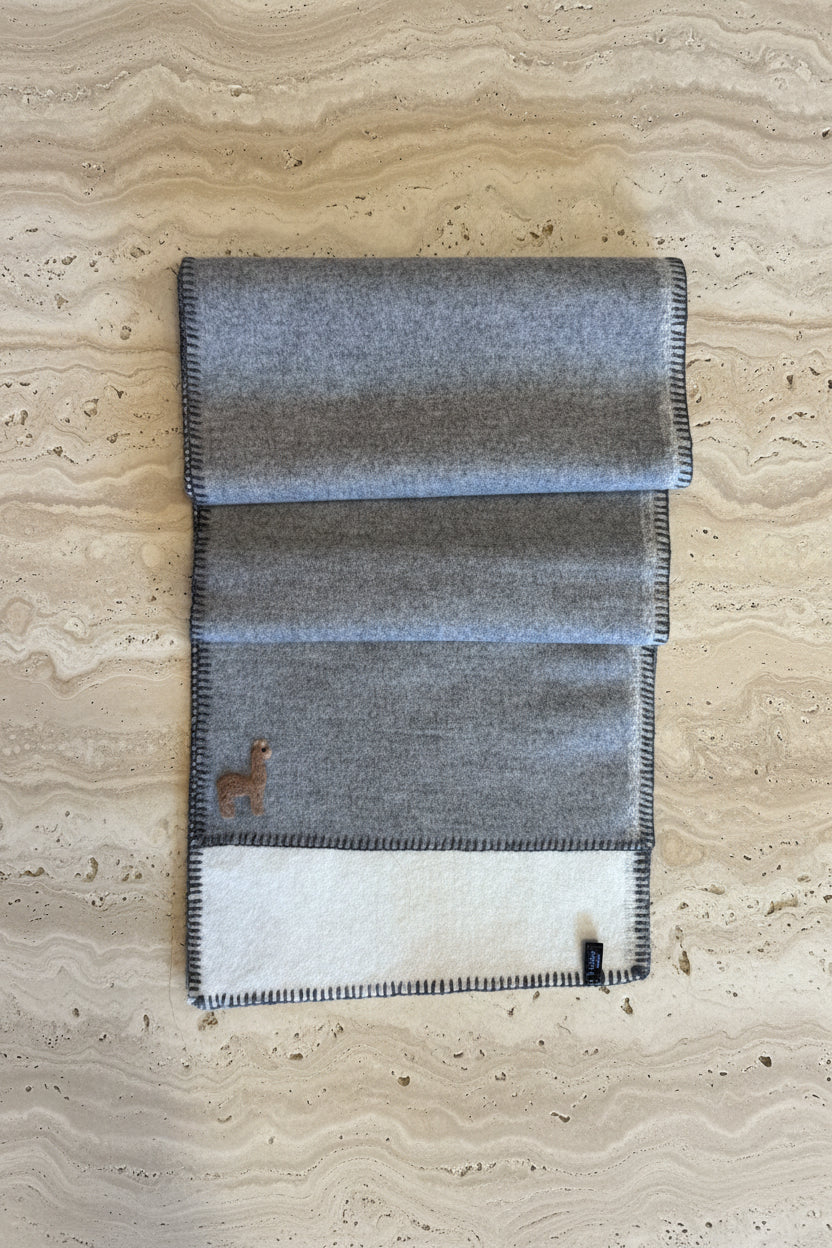 A folded gray wool blanket with black-and-white checkered fringed edges, laid flat on a warm brown wooden floor. A small embroidered deer logo is visible on the lower left.