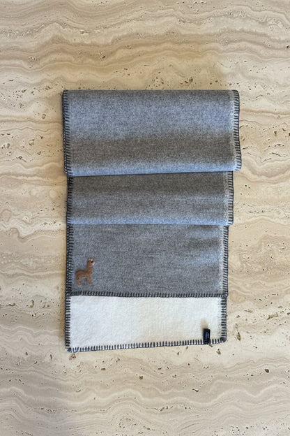 A folded gray wool blanket with black-and-white checkered fringed edges, laid flat on a warm brown wooden floor. A small embroidered deer logo is visible on the lower left.