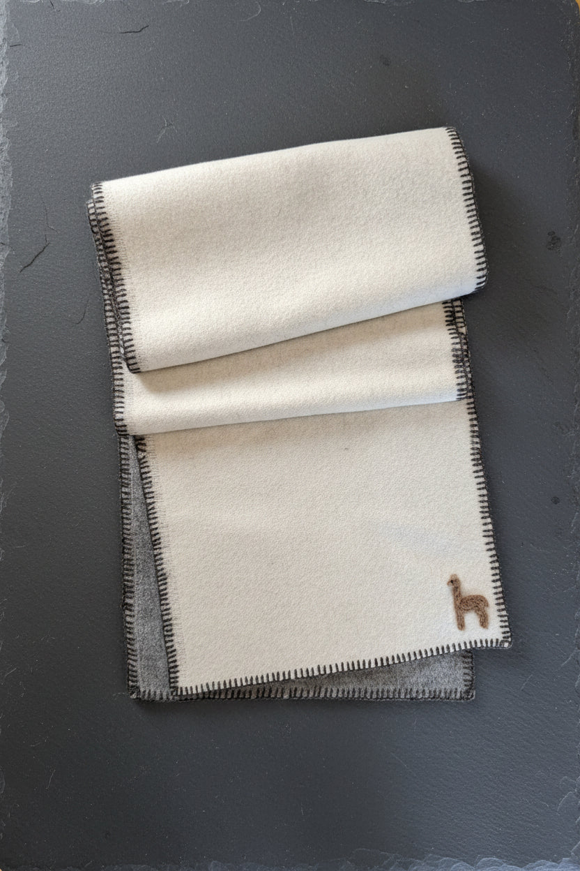 A folded cream-colored wool blanket with dark gray stitched edges and a small embroidered brown horse logo in the bottom right corner, laid flat on a wooden floor.