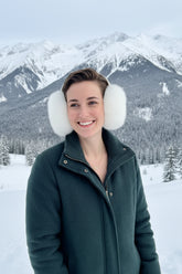 A smiling woman wearing fluffy white faux-fur earmuffs and a black, white, and pink patterned blouse, standing against a plain beige wall.