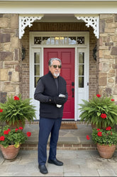A man wearing a dark gray fleece vest, black gloves, and sunglasses stands in a doorway holding a can of beer. The background features a bright red door and a striped bed.