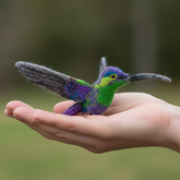 Hand holding a colorful felted hummingbird against a blurred natural background