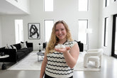 A woman with blonde hair holds three fluffy white tiny alpacas in her hand in front of a black and white living room.