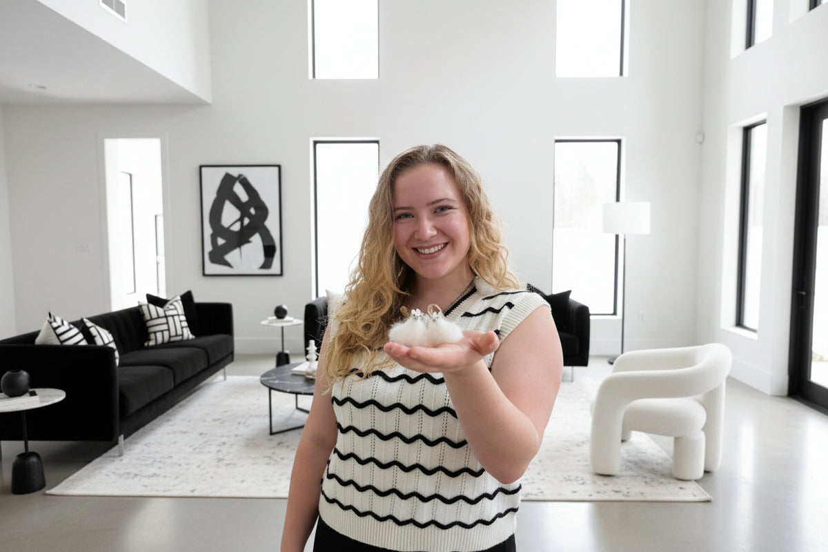 A woman with blonde hair holds three fluffy white tiny alpacas in her hand in front of a black and white living room.