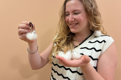 A woman with blonde hair holds a small white fluffy bunny in her hand while wearing a black and white wavy-patterned knit top. She is holding a small white fluffy bunny on a black hair tie in her other hand.