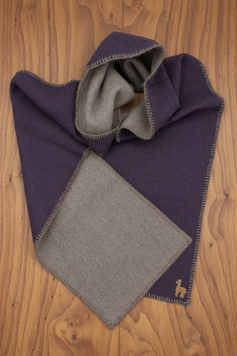 A folded dark purple wool scarf with a contrasting light brown lining and a small embroidered alpaca logo, displayed on a wood surface.