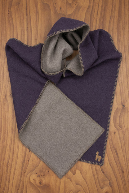 A folded dark purple wool scarf with a contrasting light brown lining and a small embroidered alpaca logo, displayed on a wood surface.