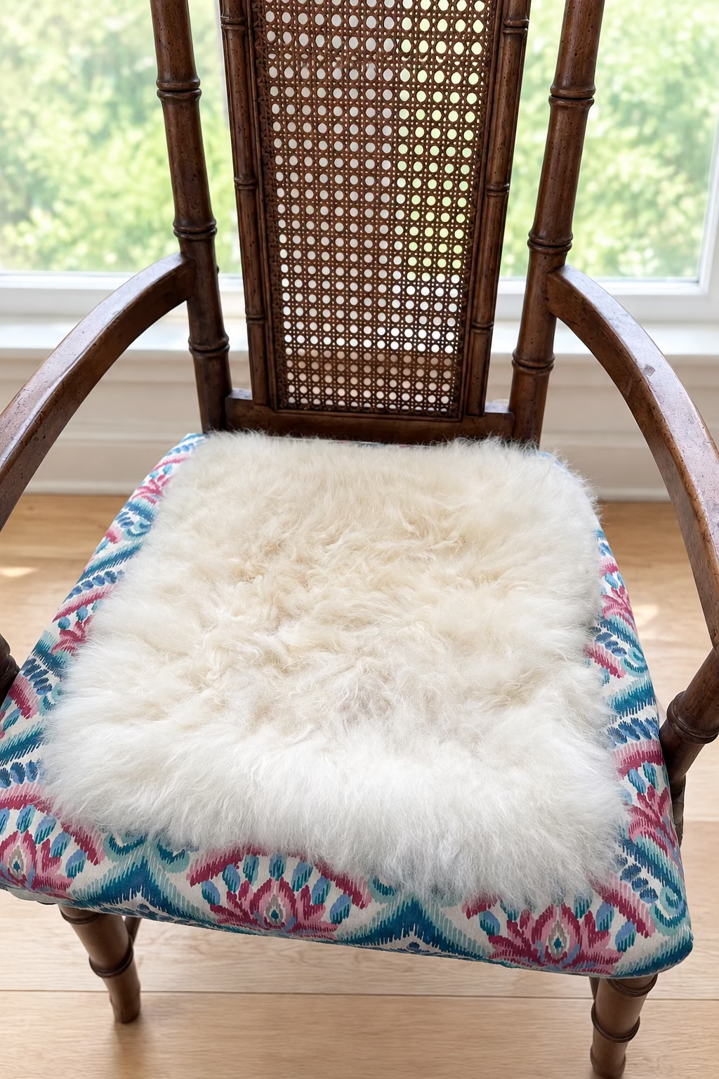 A fluffy cream-colored alpaca fur pad resting on a patterned fabric chair seat in front of a window.