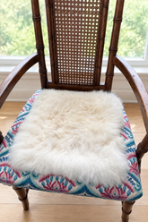 A fluffy cream-colored alpaca fur pad resting on a patterned fabric chair seat in front of a window.