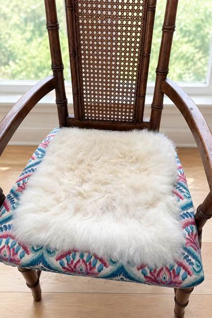 A fluffy cream-colored alpaca fur pad resting on a patterned fabric chair seat in front of a window.