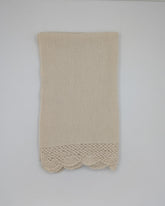 A beige alpaca shawl with a decorative crocheted lace hem, hanging on a white wall.