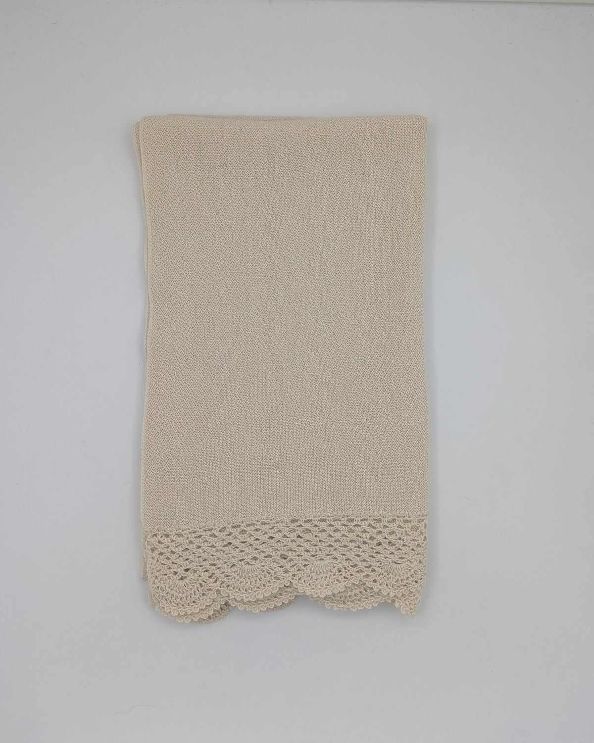 A beige alpaca shawl with a decorative crocheted lace hem, hanging on a white wall.