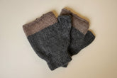 A pair of folded knit fingerless gloves in heathered brown and dark gray, with a white circular brand tag attached, laid on a light off-white textured fabric background.