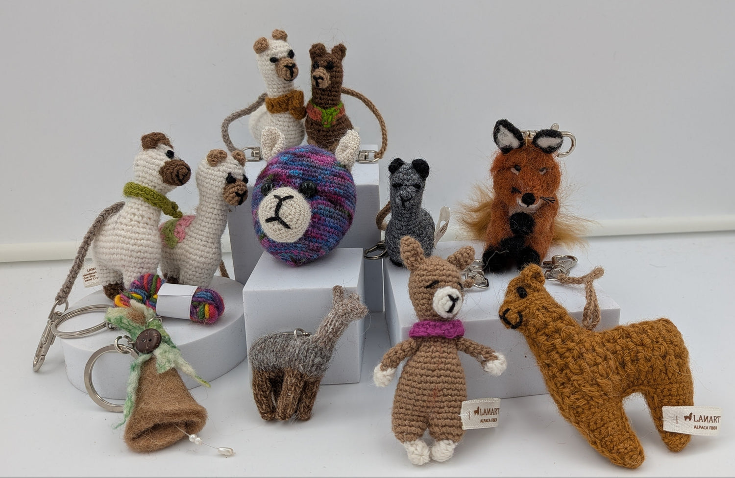 A collection of handmade crocheted animal toys displayed on white blocks against a white background. The set includes a brown llama, a gray giraffe, a brown fox, a gray cat, a white dog, a brown bear, a purple and blue ball, and other small stuffed animals.