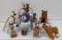 A collection of handmade crocheted animal toys displayed on white blocks against a white background. The set includes a brown llama, a gray giraffe, a brown fox, a gray cat, a white dog, a brown bear, a purple and blue ball, and other small stuffed animals.