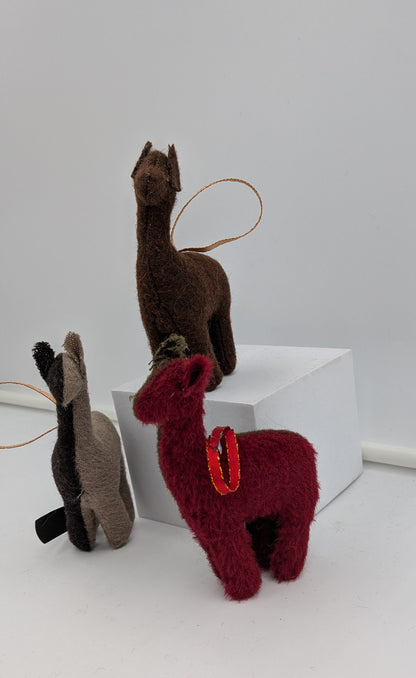 Three small alpaca or llama-shaped plush ornaments in brown, dark gray, and burgundy red, displayed on a white surface with a white block. The burgundy ornament has a small red saddle detail, and all have hanging loops.