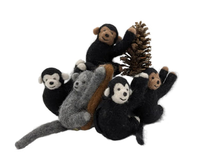 A small felted wool monkey figurine set on a white surface against a white background, featuring several black-and-white monkeys, a gray koala, and a brown pinecone.