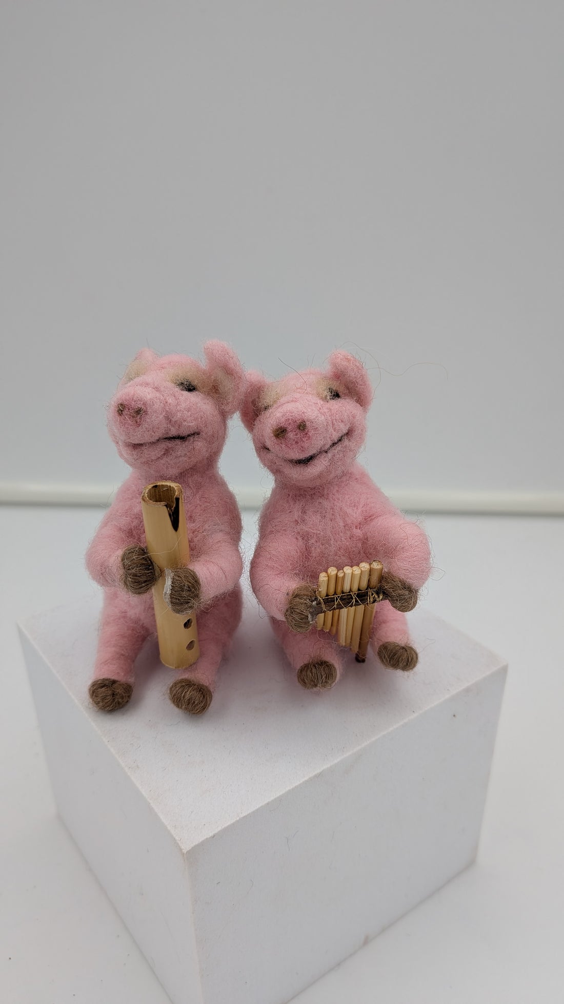 Two small pink felt pig figurines sitting on a white pedestal, one holding a gold flute-like instrument and the other holding a gold panpipe.